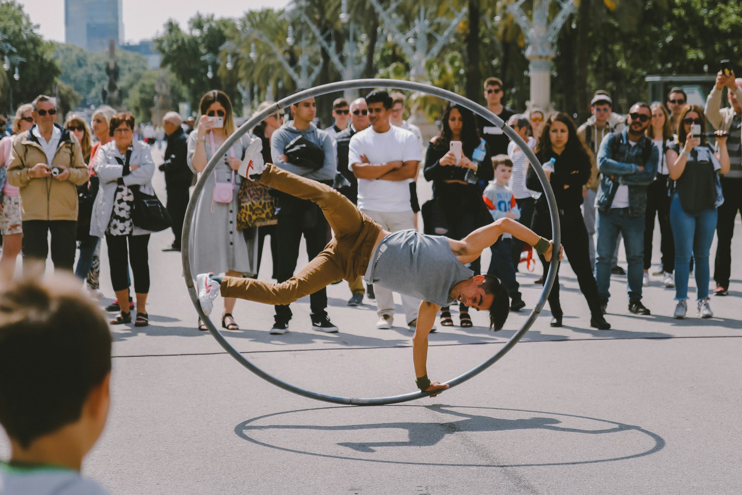 A street performer balancing inside a large steel Cyr wheel, mid-motion, in front of a watching crowd in a city park.