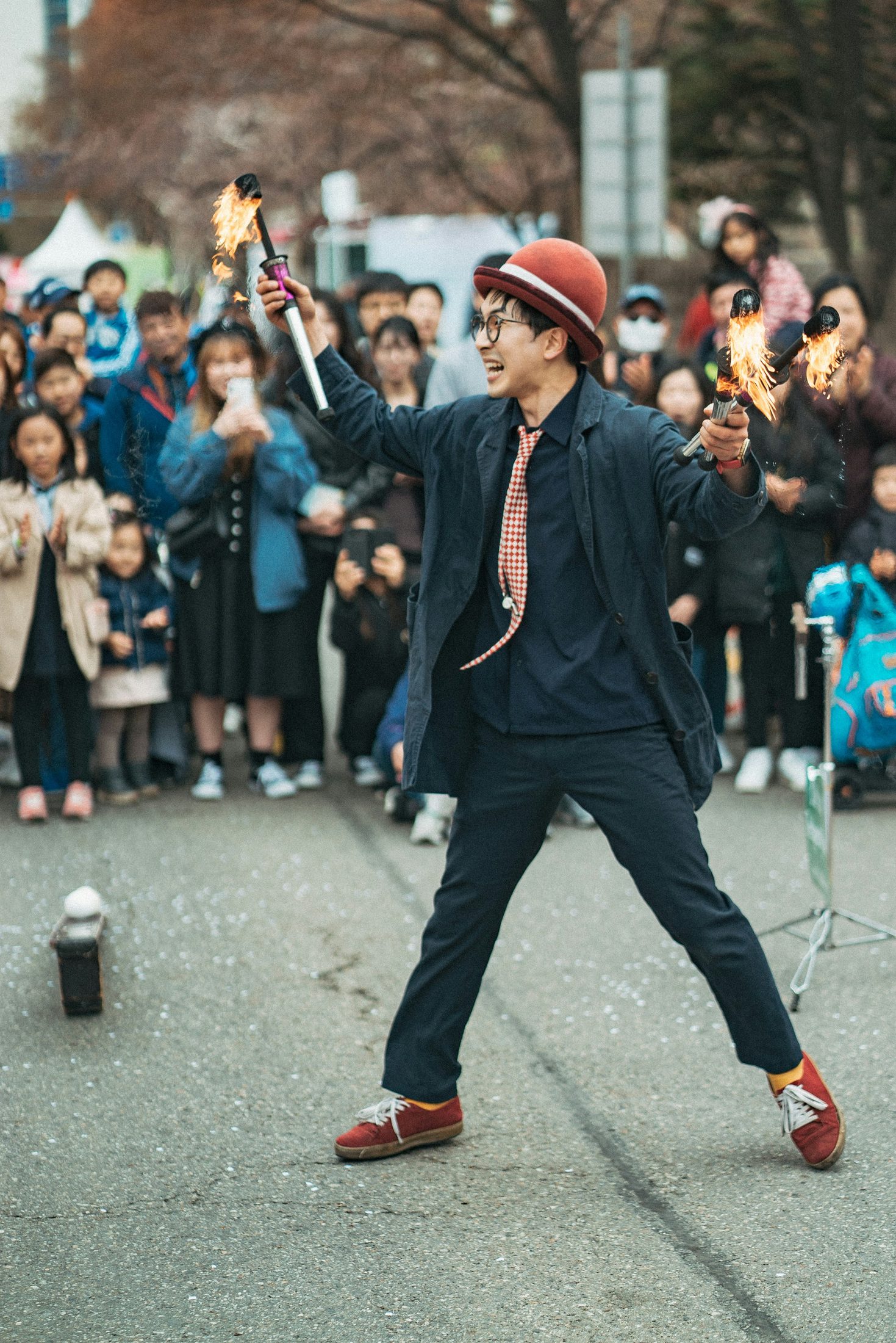 A street performer in a bowler hat swinging two flaming torches in front of an applauding crowd at dusk.