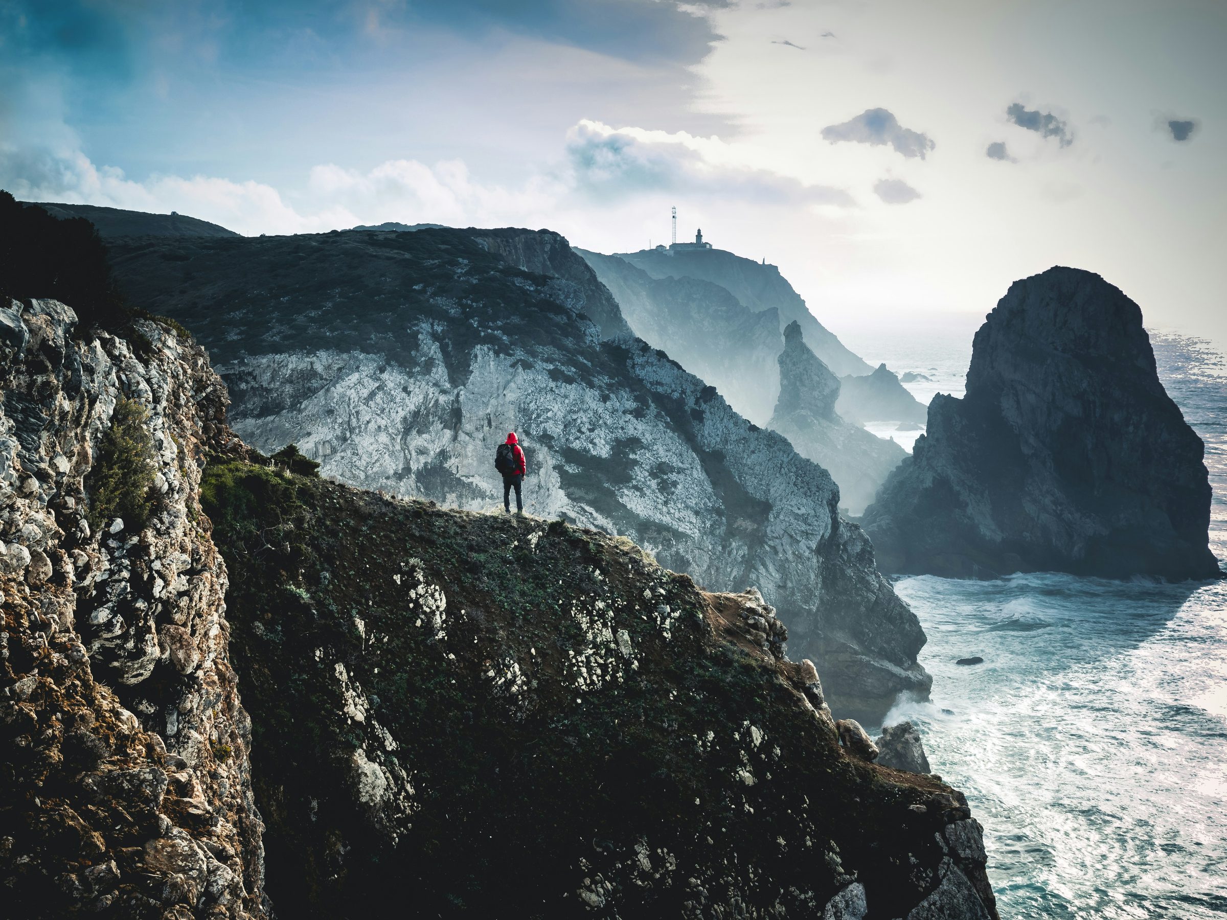 A hiker in a red jacket standing alone at the edge of a dramatic sea cliff, looking out over rock stacks and misty coastline.