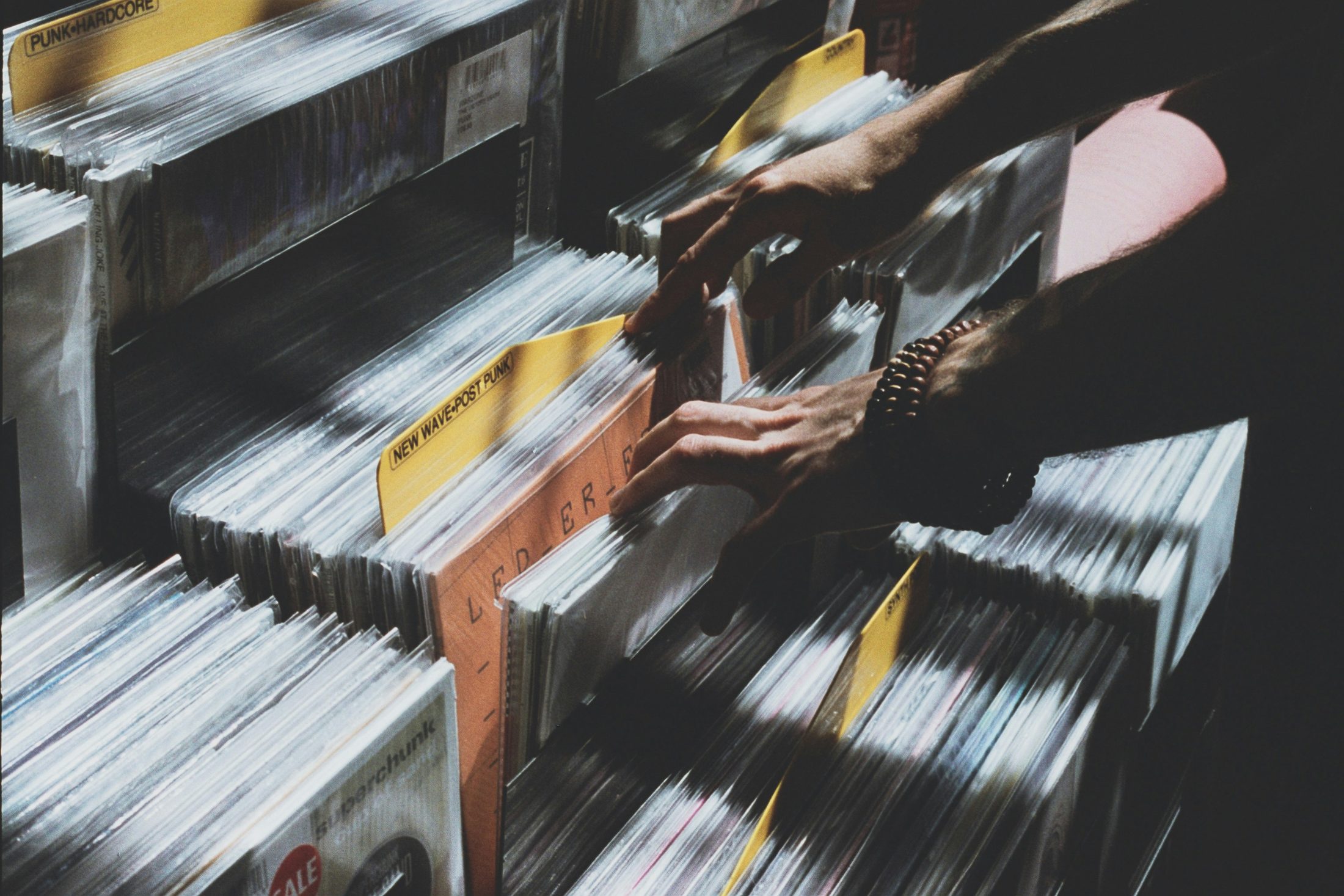 Hands flicking through vinyl records in a record store, a New Wave / Post Punk divider card visible among the sleeves.