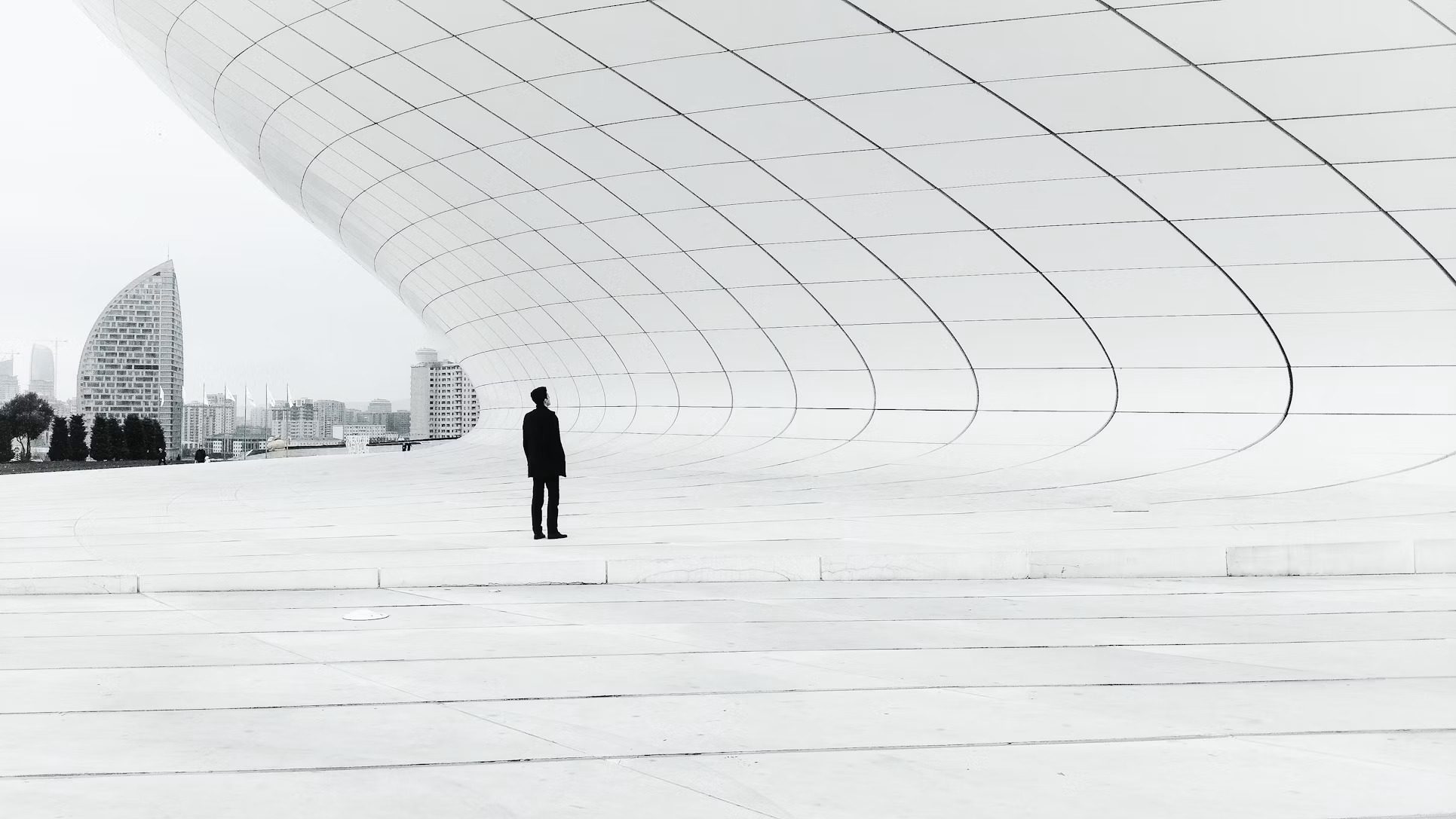 A single figure standing beneath the vast, curved white ceiling of an architectural space, dwarfed by its scale, with a distant city skyline beyond.