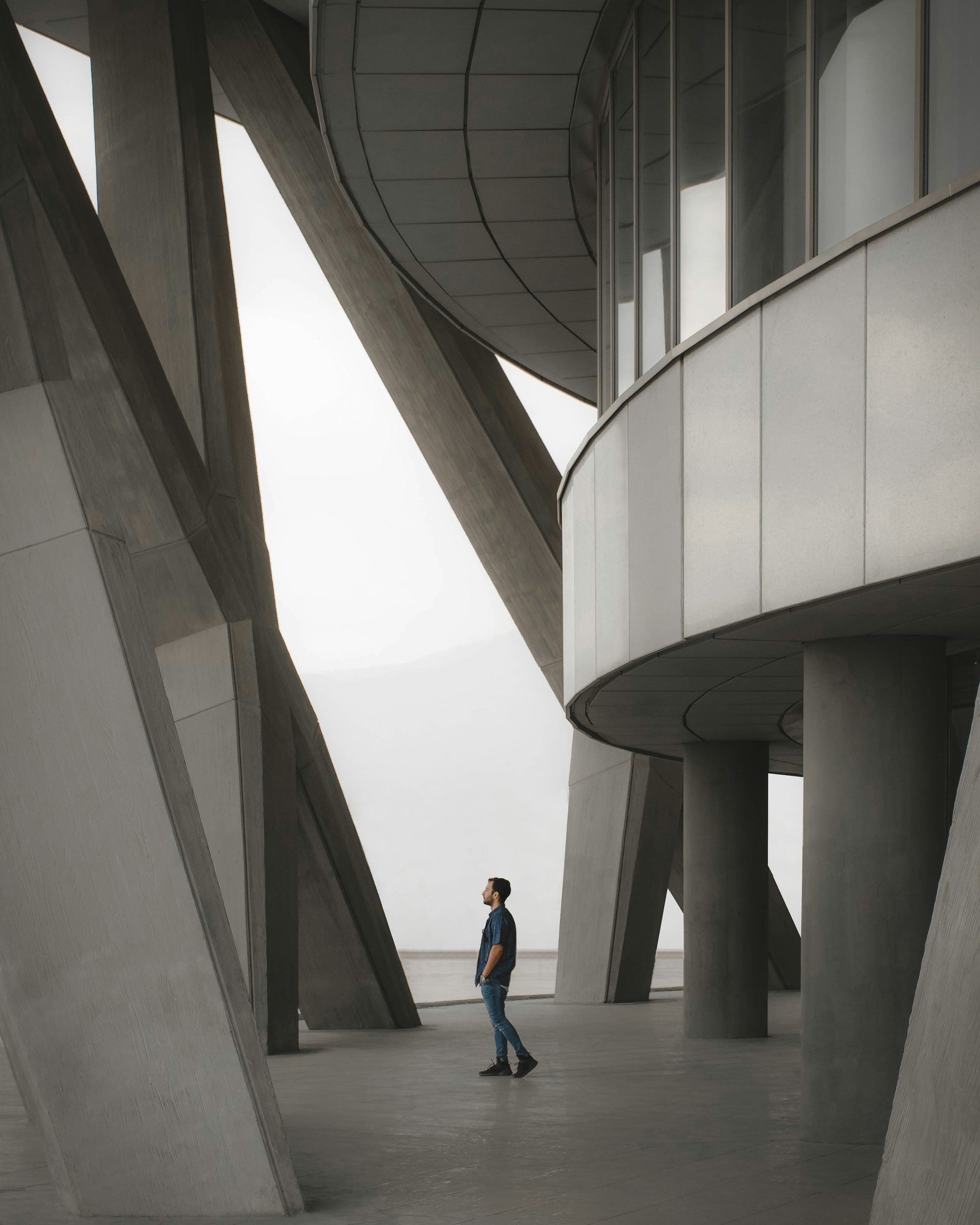 A person walking alone through a monumental concrete building, dwarfed by angled columns and a sweeping curved overhang.